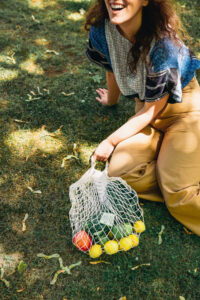 Woman sitting on grass smiling while holding a cotton bag filled with fruit.
