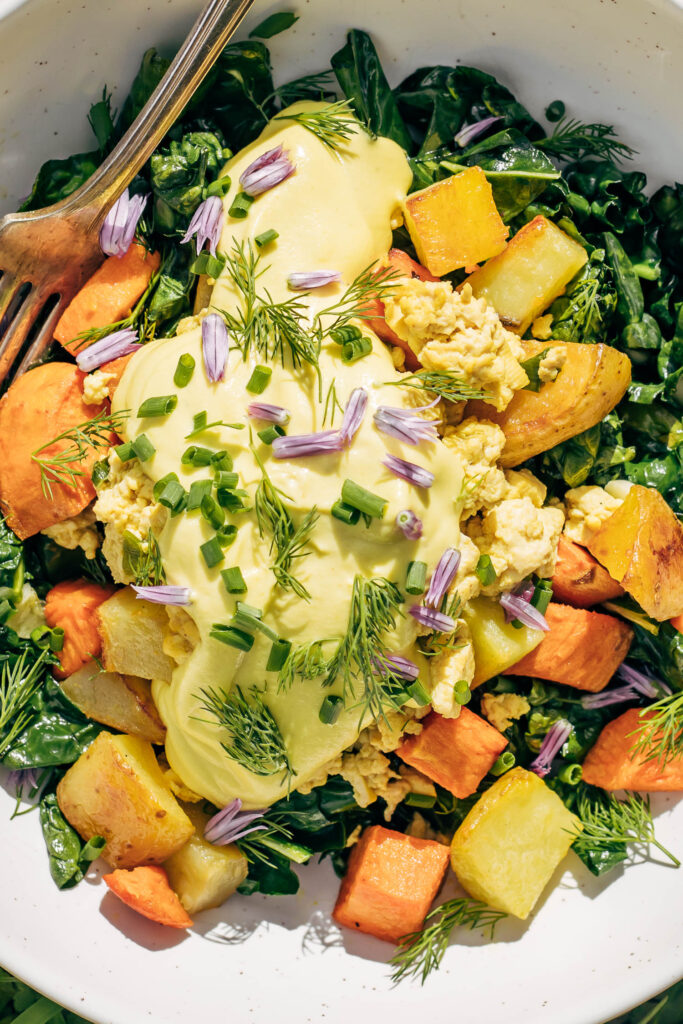 Close up of golden vegan hollandaise  made with cashews on a breakfast bowl with herbs, scramble, and yams