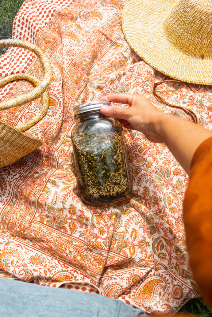 Jar of herbs on a pink picnic blanket along with basket and straw hat.