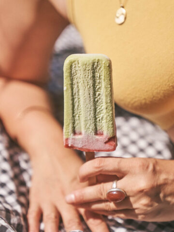 A hand holding a green matcha popsicle with a picnic background behind
