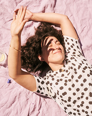 A woman lying on a blanket outdoors shading her eyes with a hand.