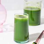 Green juice in a glass with a pitcher of juice in background.