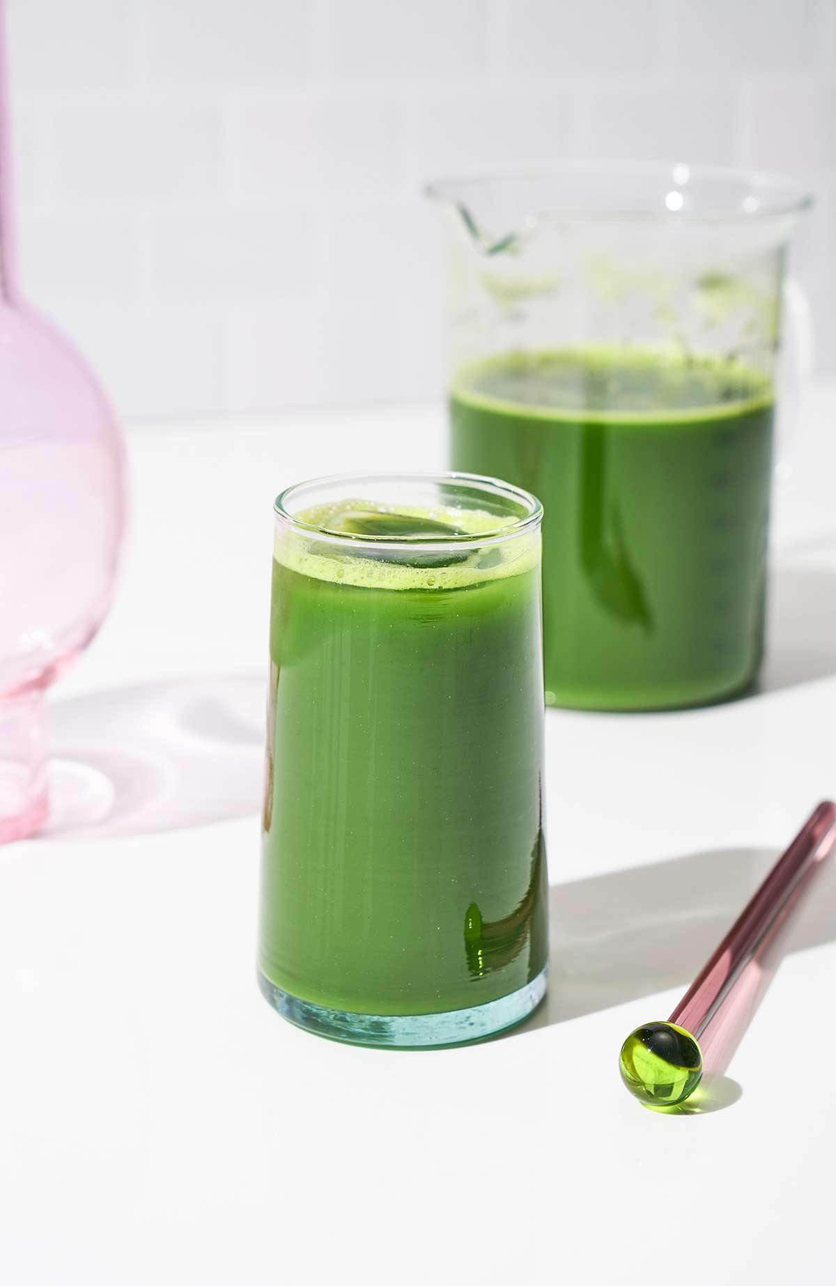 Green juice in a glass with a pitcher of juice in background.