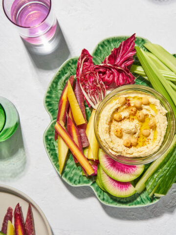 Hummus in a bowl on a plate surrounded by vegetables and drinks.