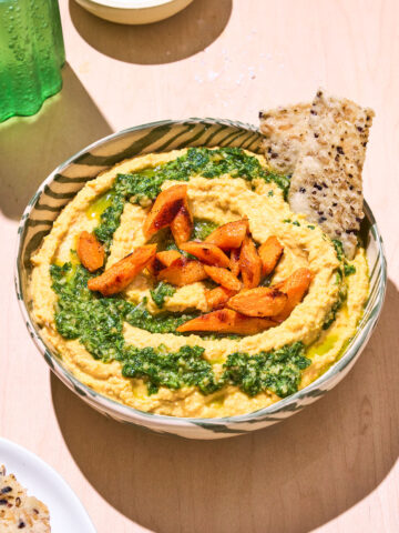Hummus in a bowl topped with pesto and roasted carrot pieces.