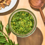 Top-down view of a jar of pesto with parsley and walnuts around.