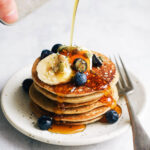 A stack of pancakes topped with fruit, with syrup being poured on.
