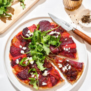 A beetroot tart topped with arugula and crumbly cheese, one slice cut.