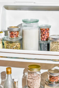 Several glass jars in a fridge door, a big one with milk and several with nuts and seeds.