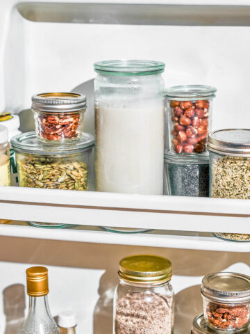 Several glass jars in a fridge door, a big one with milk and several with nuts and seeds.