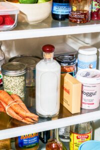 A large glass bottle filled with milk in the fridge with containers and vegetables.