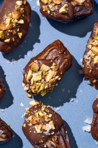 Close up of filled dates dipped in chocolate with pistachios on a blue plate.